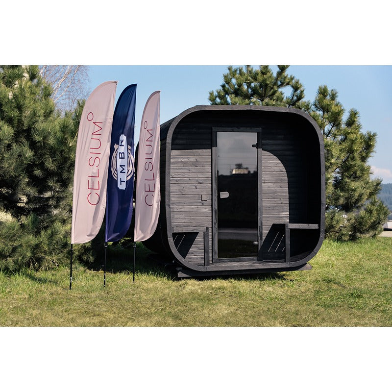 Front view of the black Celsium Ula Large sauna outdoors next to three event flags against a green backdrop